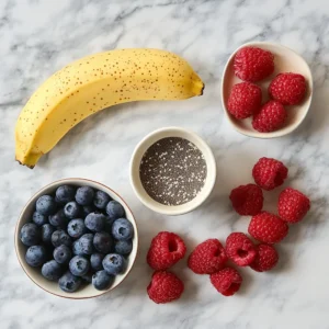 A variety of ingredients for a banana berry protein smoothie arranged on a clean counter, including a banana, mixed berries, protein powder, almond milk, and chia seeds.